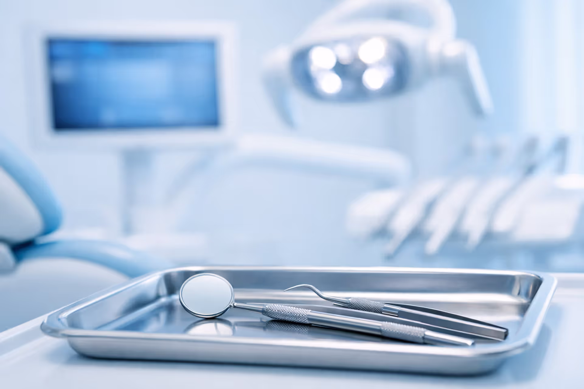 Modern dental office with a dental chair and a tray of clean dental instruments including a mirror, probe, and tweezers in a bright blue and white clinical setting