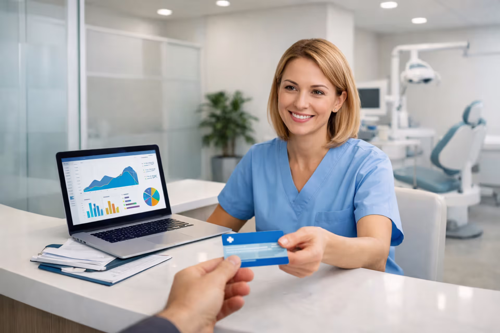 Dental office front desk administrator receiving insurance card from patient with laptop and paperwork on desk